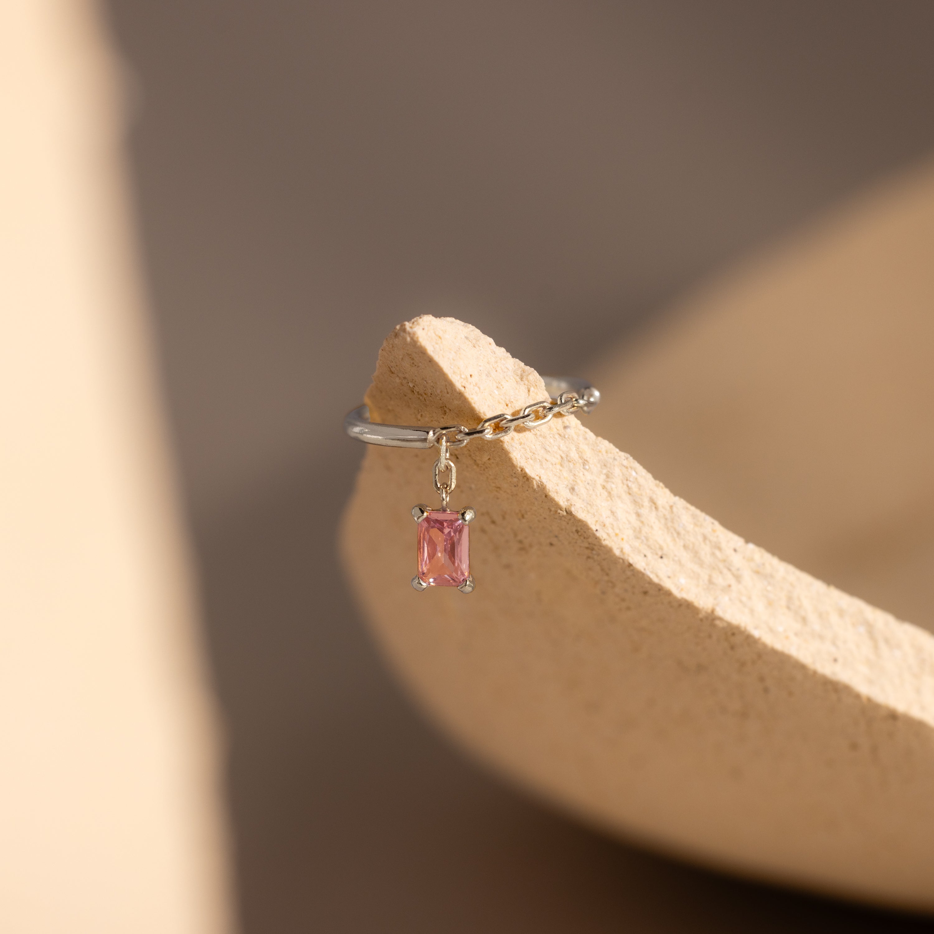 The Pink Tourmaline Drop Chain Ring features a silver chain design with a dangling pink tourmaline gemstone, displayed on a textured beige surface for a delicate, modern look.