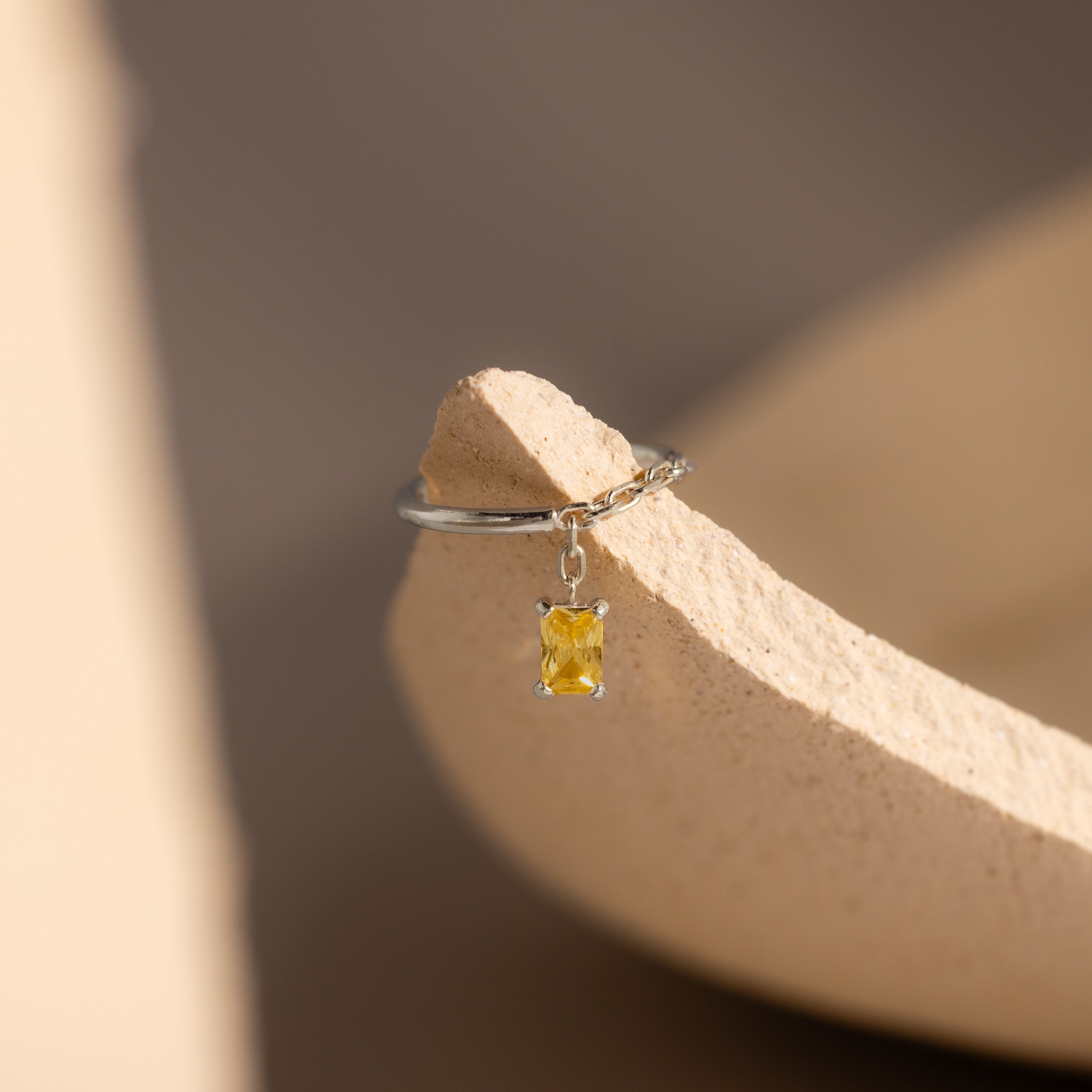 The Topaz Drop Chain Ring features a silver chain band with a small emerald-cut yellow topaz pendant, elegantly displayed on a textured beige surface.