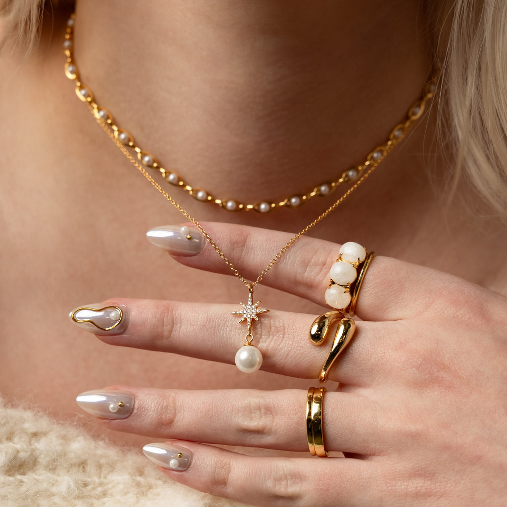 A womans hand with gold rings, pearl accents, layered necklaces featuring the North Star Pearl Necklace, and pearl-adorned nails.