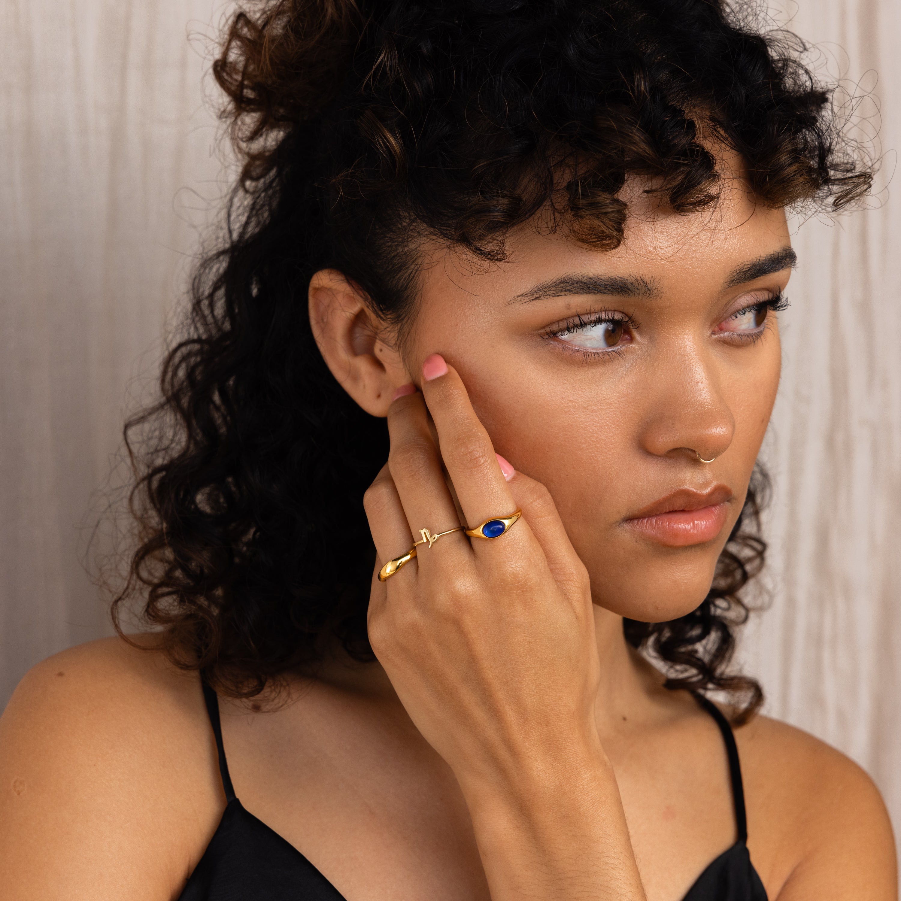 A woman with curly hair in a black top touches her face, wearing the Bold Mood Ring in gold, and looks to the side.