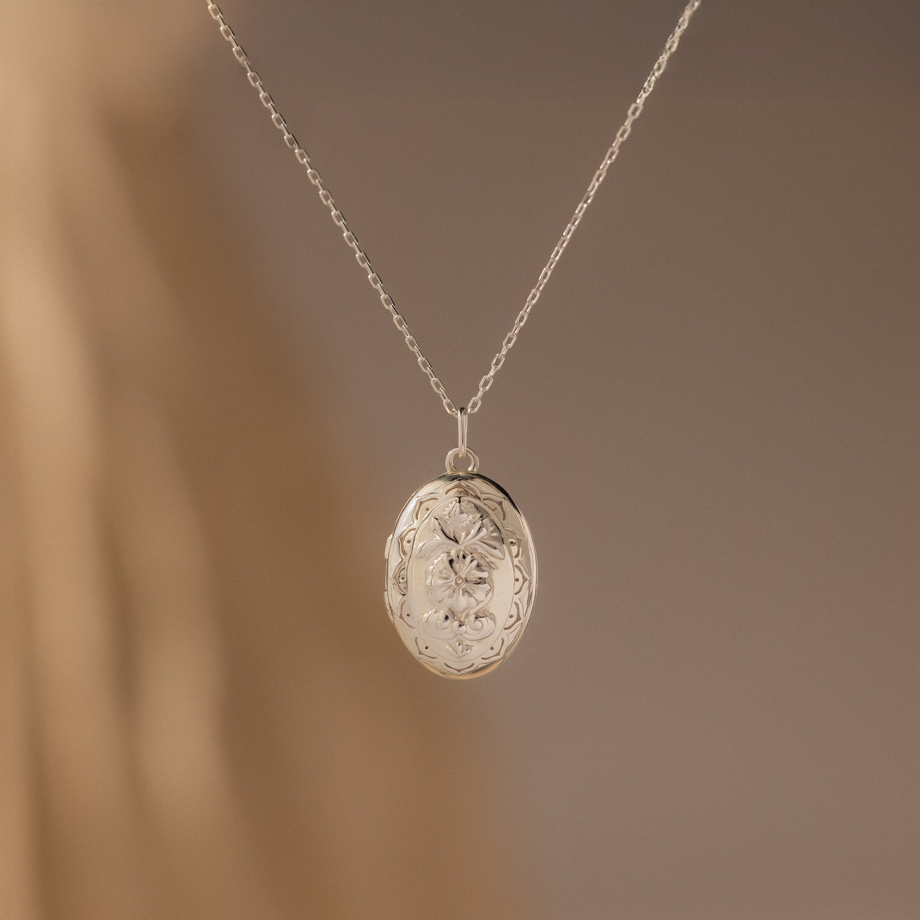 The Floral Handwriting Locket Necklace features an oval silver pendant with delicate engravings, hanging from a fine chain and set against a softly blurred neutral background.