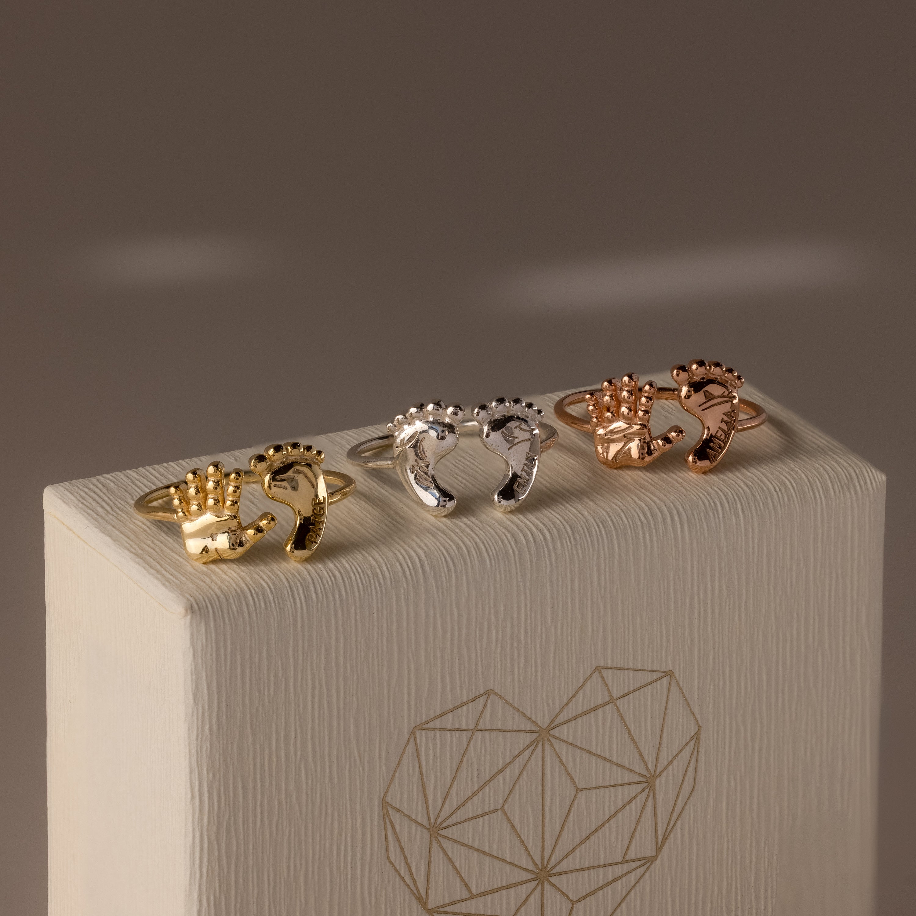 The Hand & Footprint Duo Name Ring in Sterling Silver, adorned with hand and foot charms, is beautifully displayed on a textured box—an ideal personalized keepsake gift for new parents.