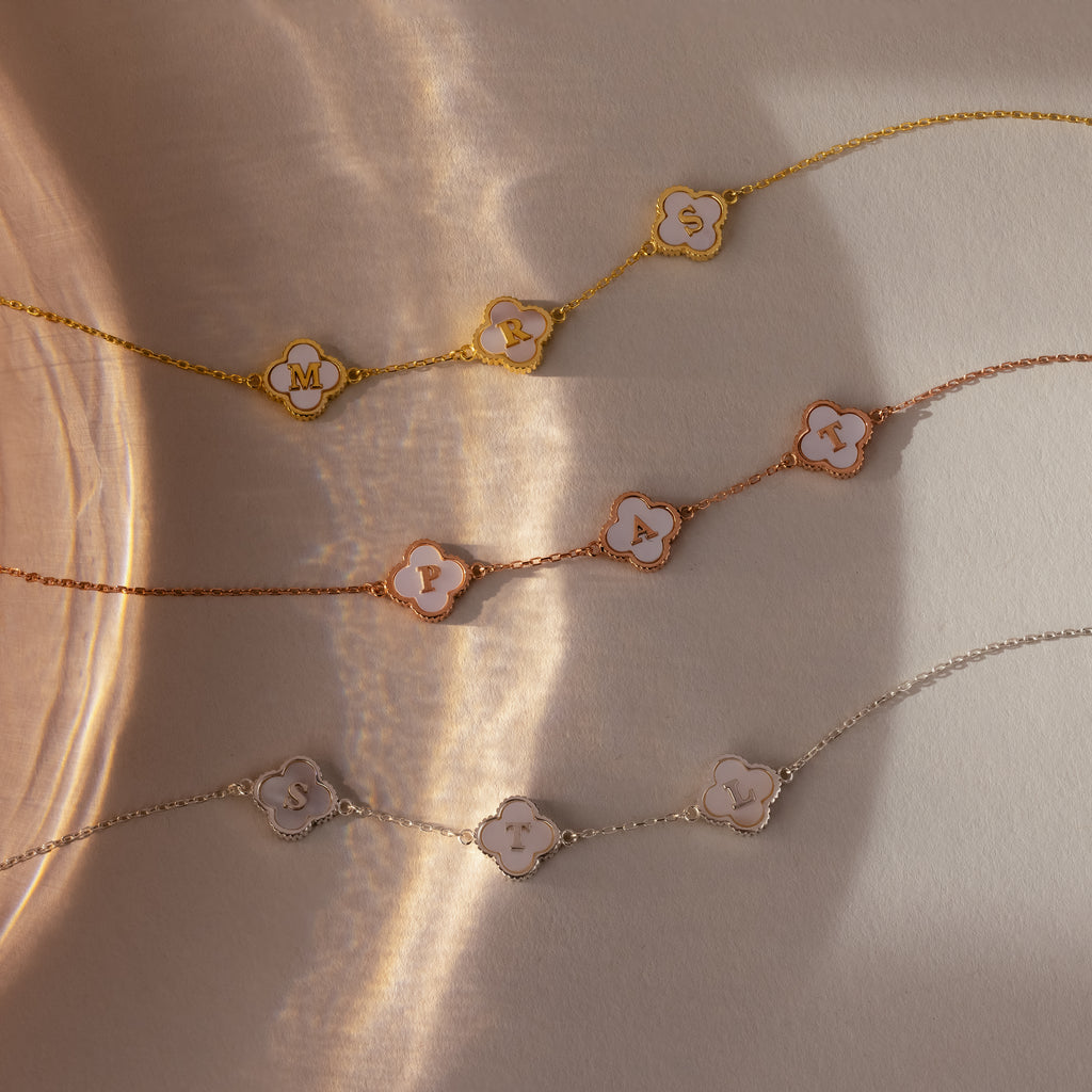The Pearl Clover Letter Bracelet features delicate chain links, an initial letter charm, and a mother of pearl clover, all showcased on a neutral, sunlit background.