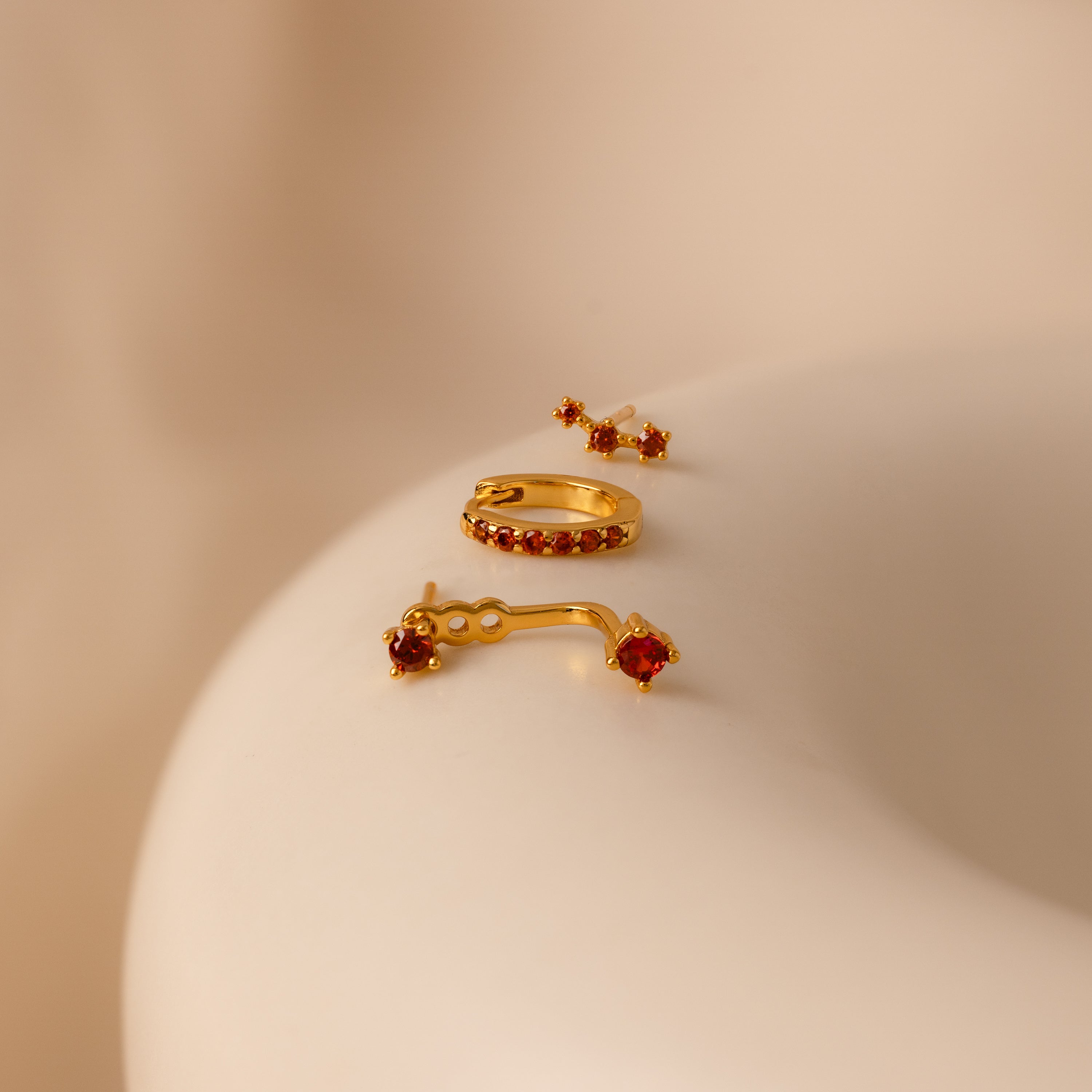 The Alexandrite Earring Trio Set features three gold earrings with red gemstones in elegant settings, including a pavé huggie hoop, displayed on a smooth beige surface.
