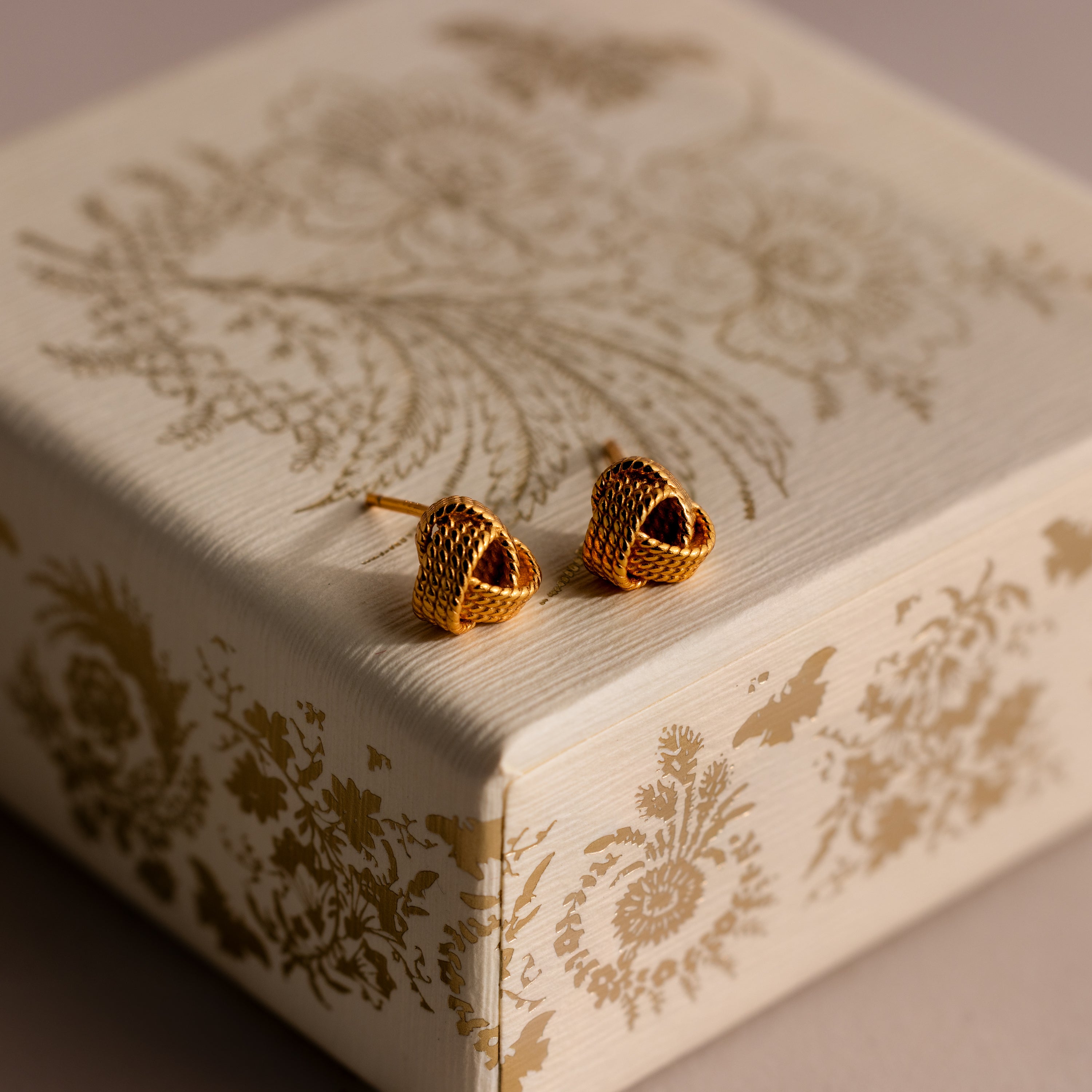 Rope Knot Studs in 18K Gold displayed on an ornate cream and gold patterned box.
