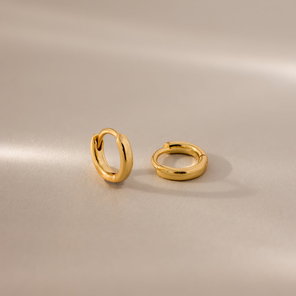 Pair of 6mm tiny dainty gold huggie hoop earrings displayed on a neutral satin surface with soft lighting.