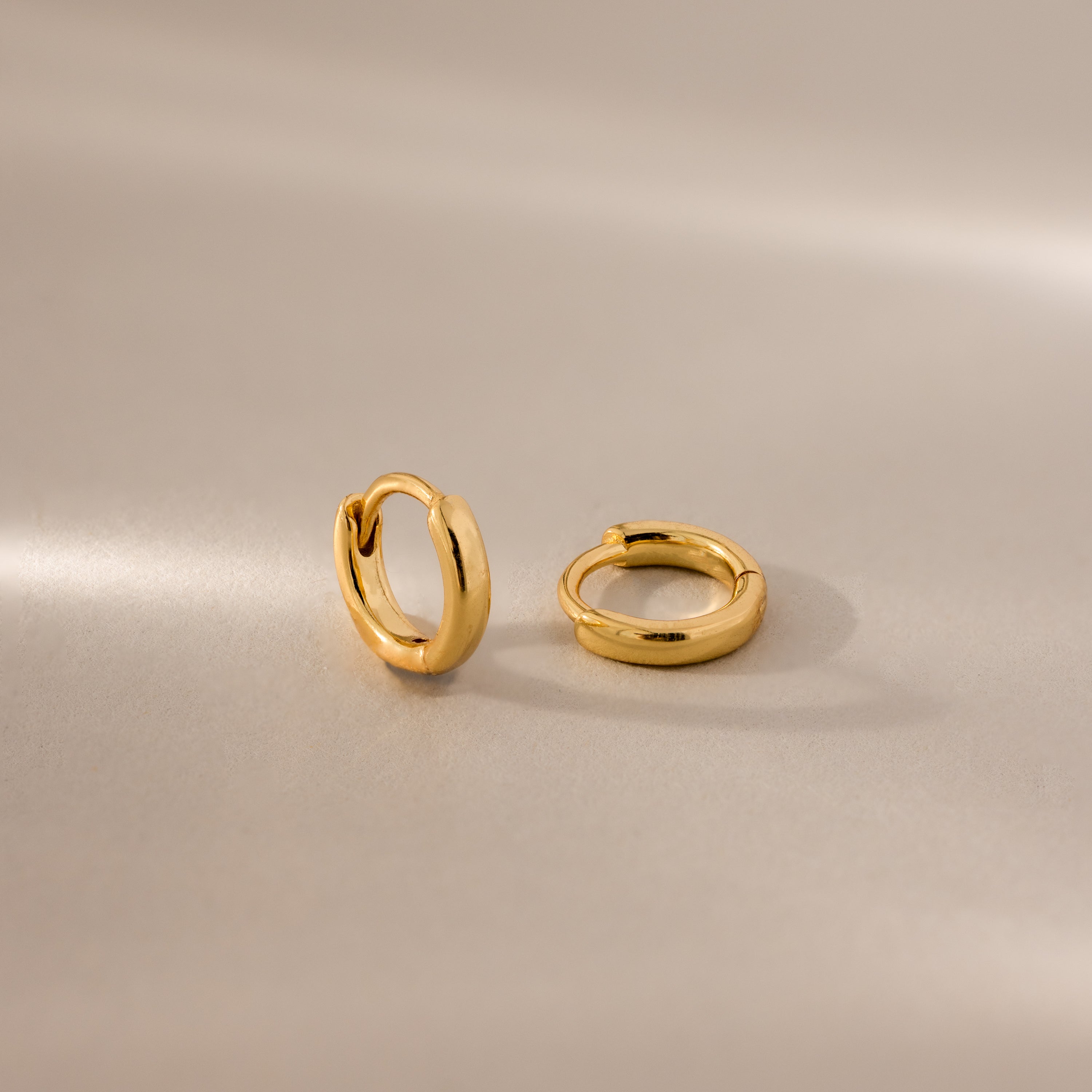 Pair of 6mm tiny dainty gold huggie hoop earrings displayed on a neutral satin surface with soft lighting.