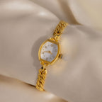 The Heirloom Chain Link Timepiece, featuring a gold chain band, white face, and diamond accents, is elegantly displayed on cream satin fabric.