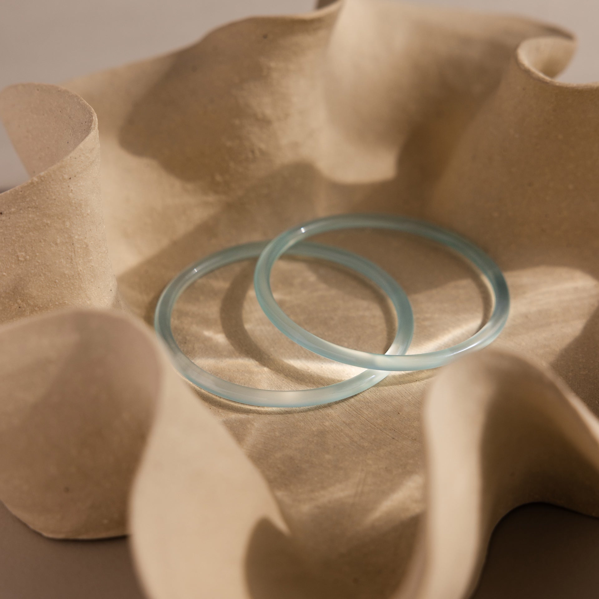 Two thin blue jade bangles from the Thin Blue Jade Bangle Set are displayed in a wavy, textured beige ceramic bowl.