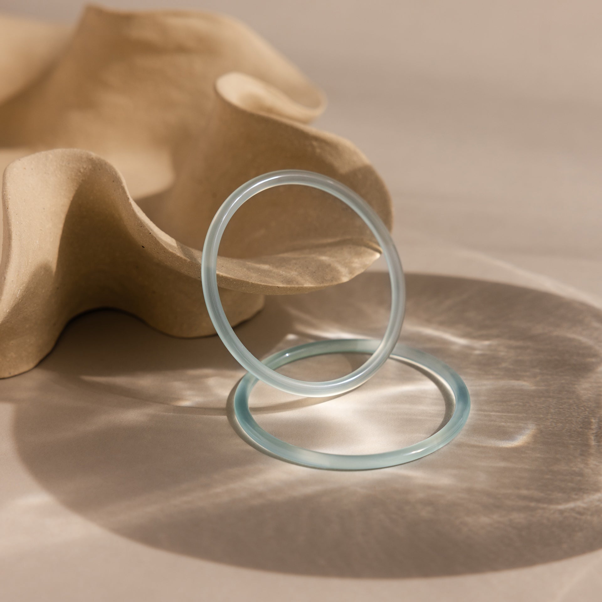 The Thin Blue Jade Bangle Set, featuring two translucent bands, is displayed on a beige surface next to a wavy sculptural object.