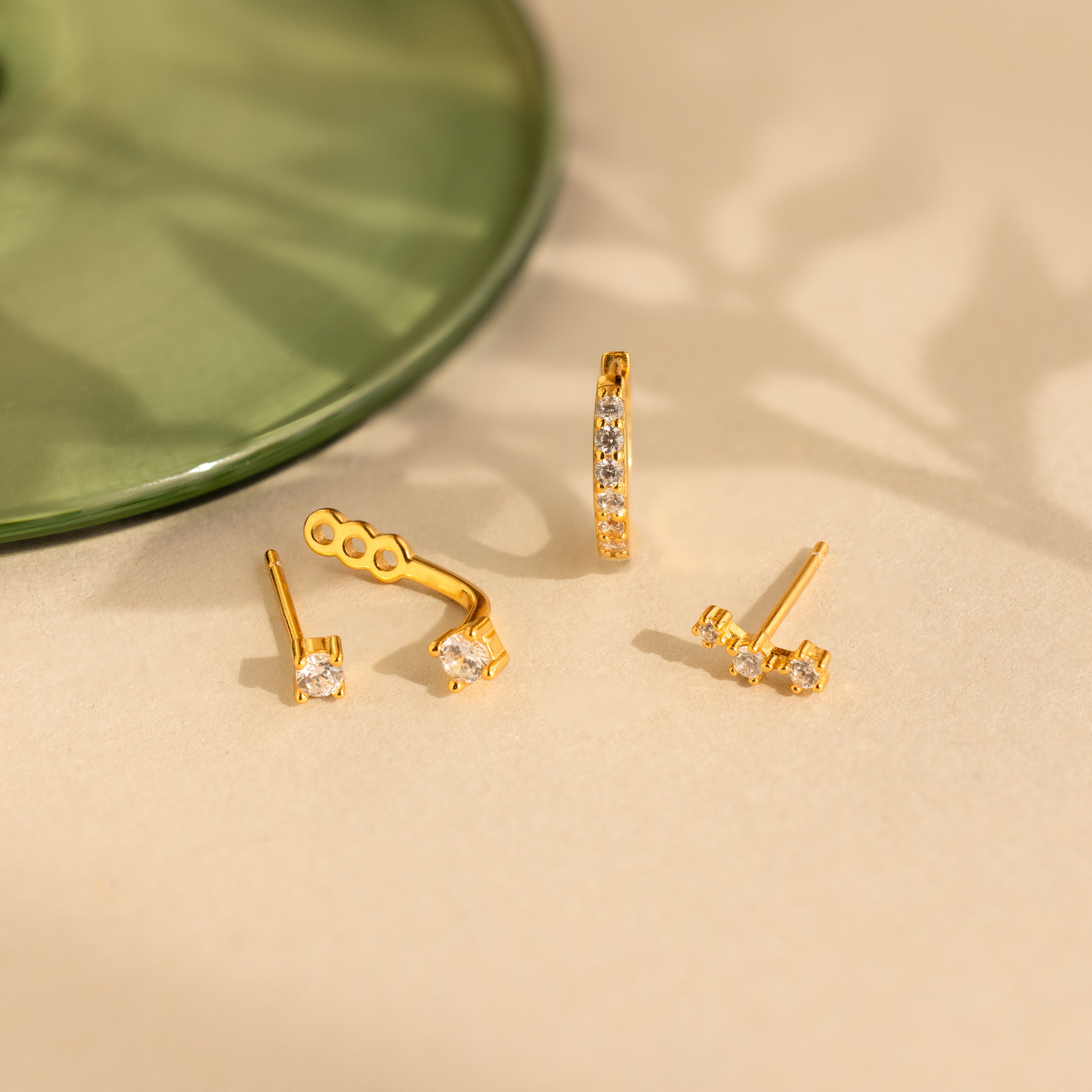 Three pairs of gold and diamond earrings, including a Diamond Earrings Set in 18K Gold and Pave Huggie Hoops, are displayed on a beige surface with a green glass object nearby.