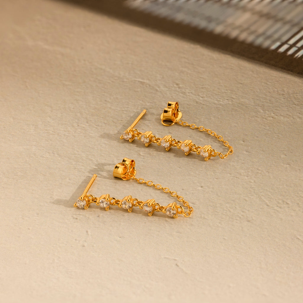 The Diamond Chain Drop Earrings feature a row of sparkling stones dangling elegantly from a delicate chain against a beige textured surface.