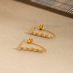 The Diamond Chain Drop Earrings feature a row of sparkling stones dangling elegantly from a delicate chain against a beige textured surface.