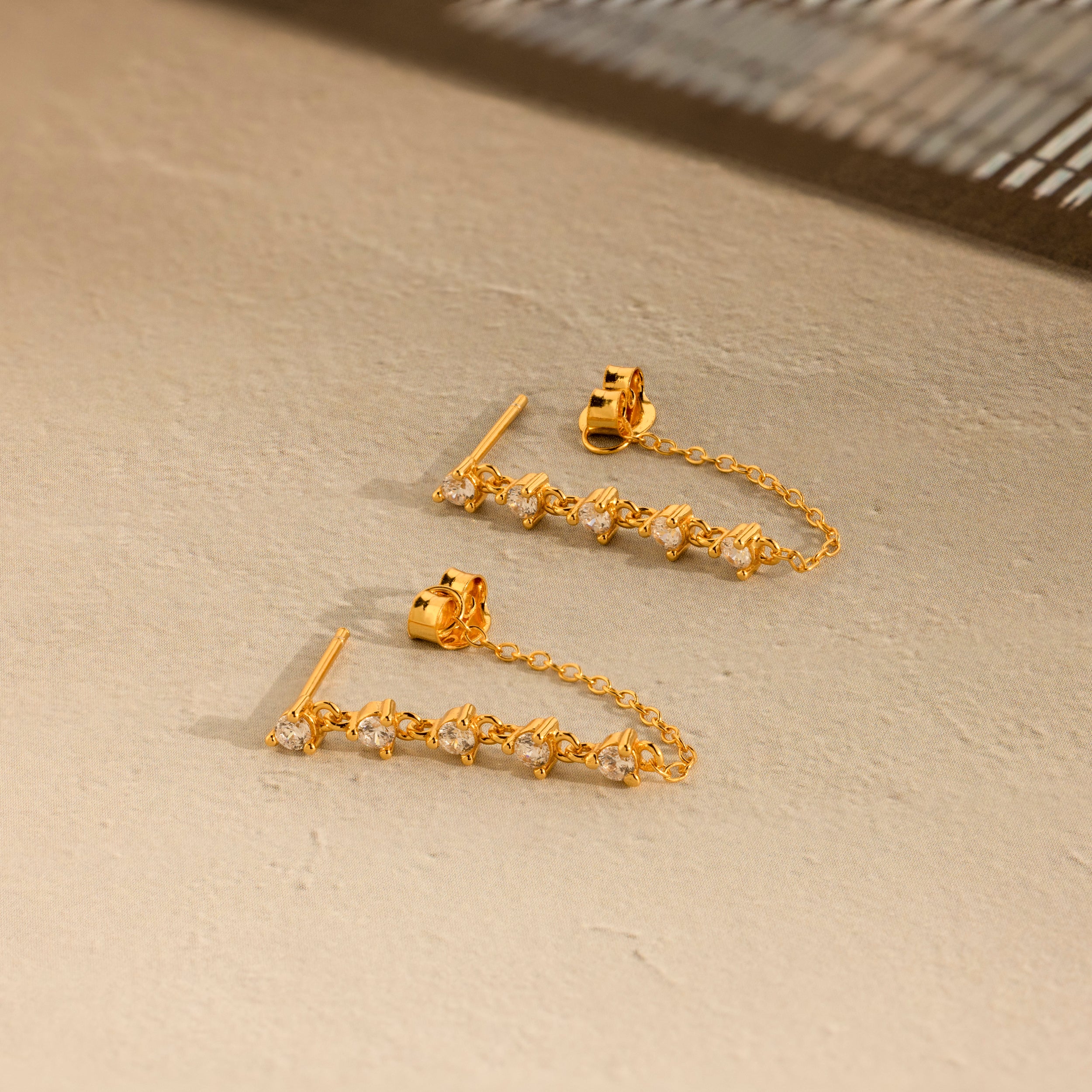 The Diamond Chain Drop Earrings feature a row of sparkling stones dangling elegantly from a delicate chain against a beige textured surface.