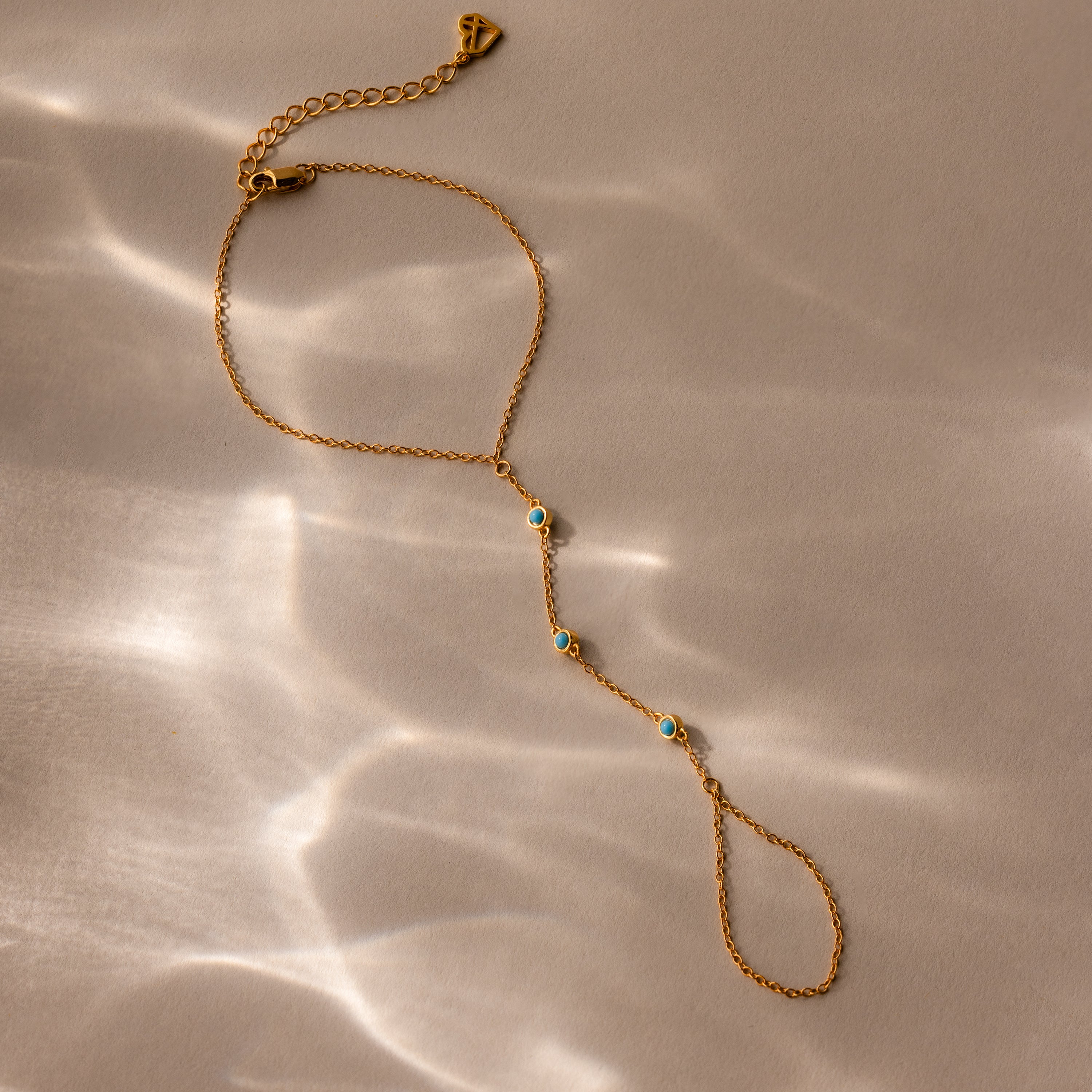 The Turquoise Hand Chain Bracelet features three vibrant stones on a gold chain, displayed on a beige surface and illuminated by soft light reflections.