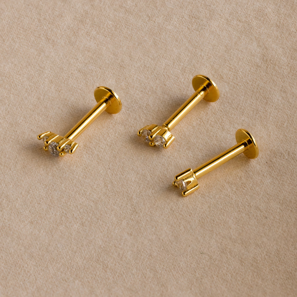 Three dainty earrings, including the Tiny Diamond Flatback Studs Set in 18K Gold, are displayed on a beige fabric background.