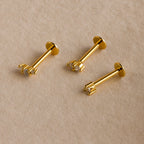 Three dainty earrings, including the Tiny Diamond Flatback Studs Set in 18K Gold, are displayed on a beige fabric background.