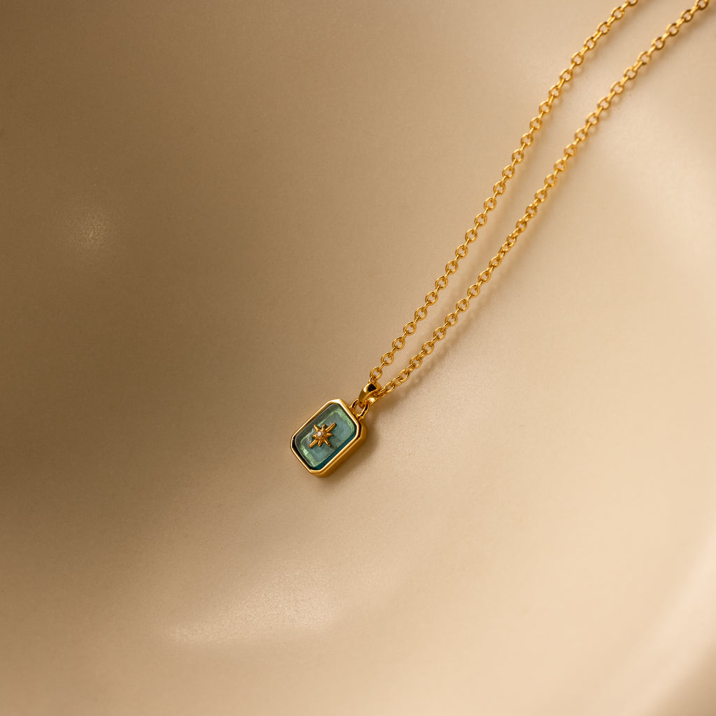 The North Star Blue Zircon Necklace features a gold chain with a rectangular blue zircon pendant and a small star, shown on a beige background.