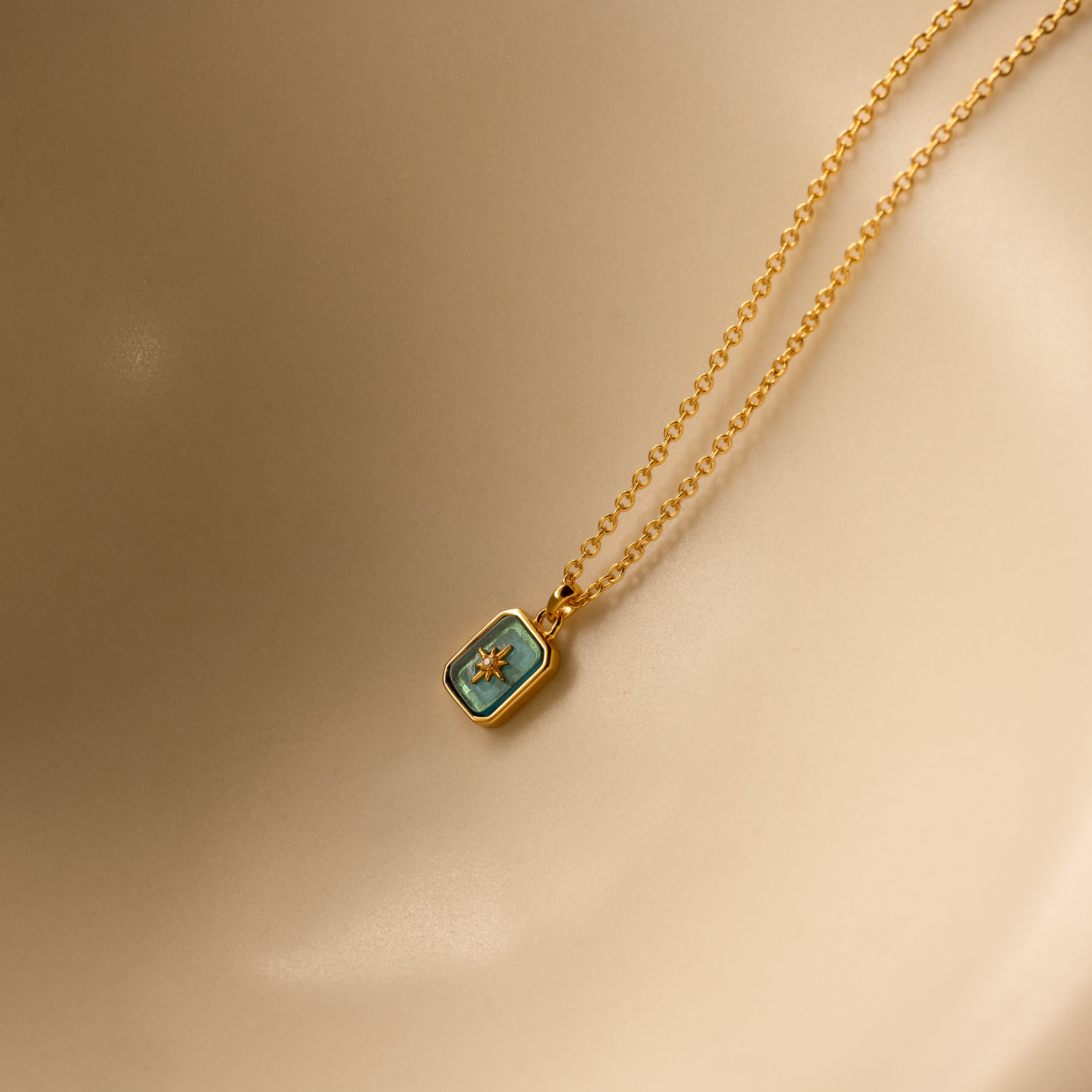 The North Star Blue Zircon Necklace features a gold chain with a rectangular blue zircon pendant and a small star, shown on a beige background.
