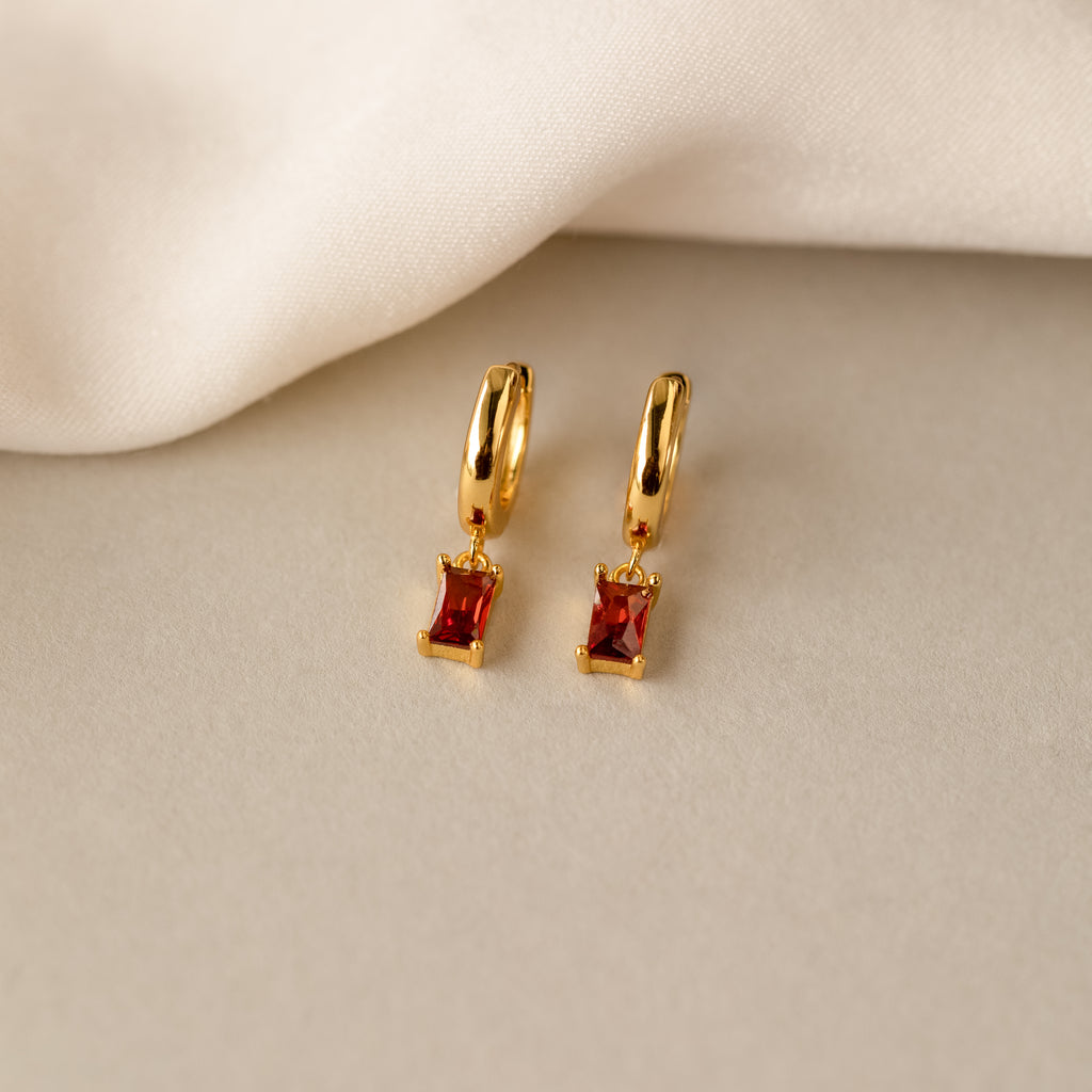 Gold huggie hoop earrings with rectangular baguette-cut garnet charms, displayed on a soft cream fabric backdrop.