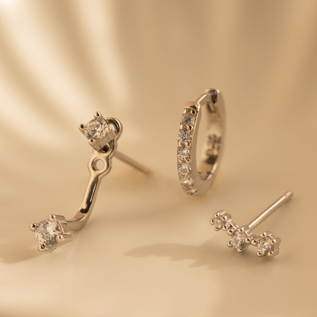 The Diamond Earrings Set in Sterling Silver features three elegant designs—including a chic Ear Climber and sparkling Pave Huggie Hoop—beautifully displayed on a smooth, cream surface, perfect for any occasion.
