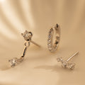 The Diamond Earrings Set in Sterling Silver features three elegant designs—including a chic Ear Climber and sparkling Pave Huggie Hoop—beautifully displayed on a smooth, cream surface, perfect for any occasion.