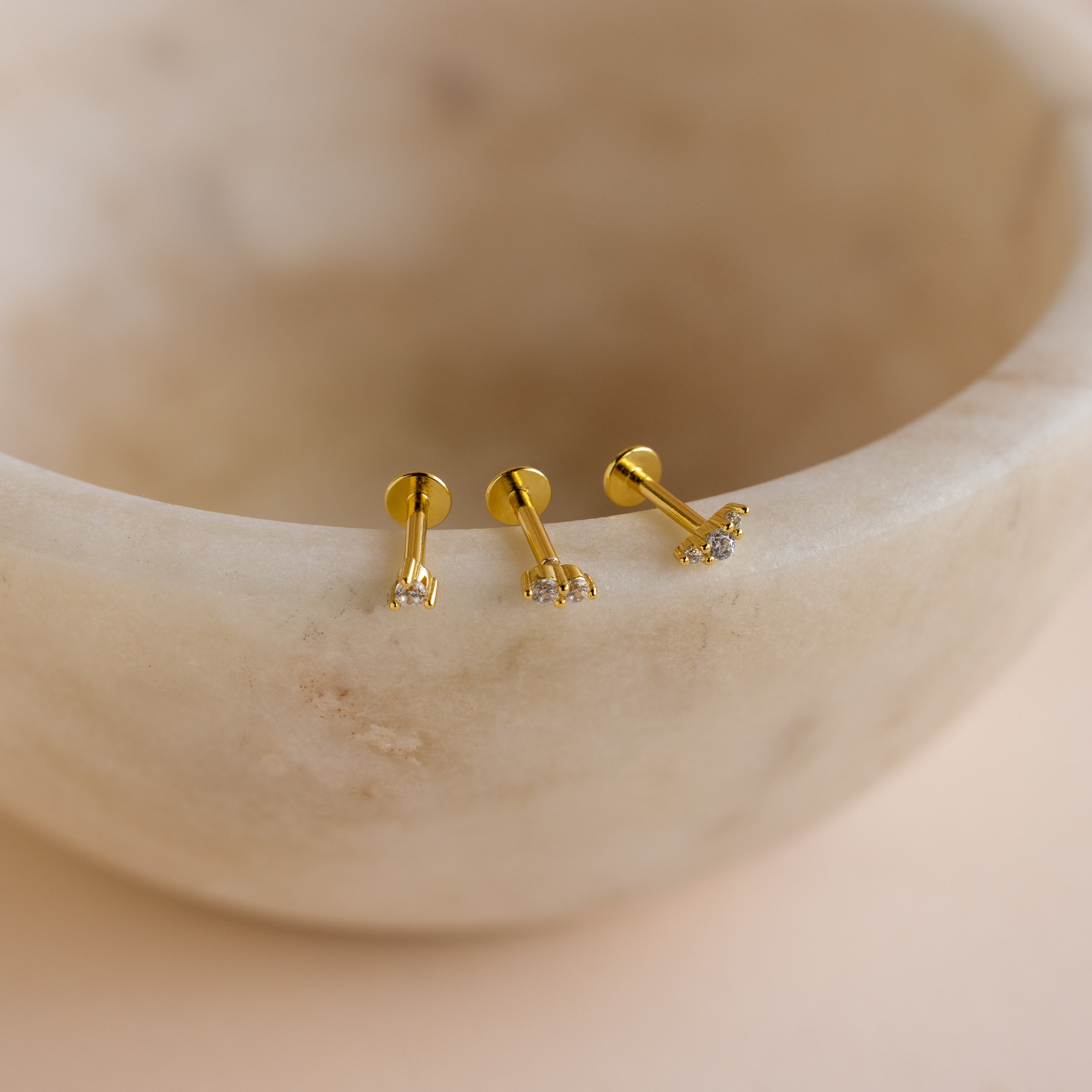 Three dainty earrings with clear stones rest on the rim of a smooth, beige marble bowl—perfect Tiny Diamond Flatback Studs Set in 18K Gold for an elegant touch.
