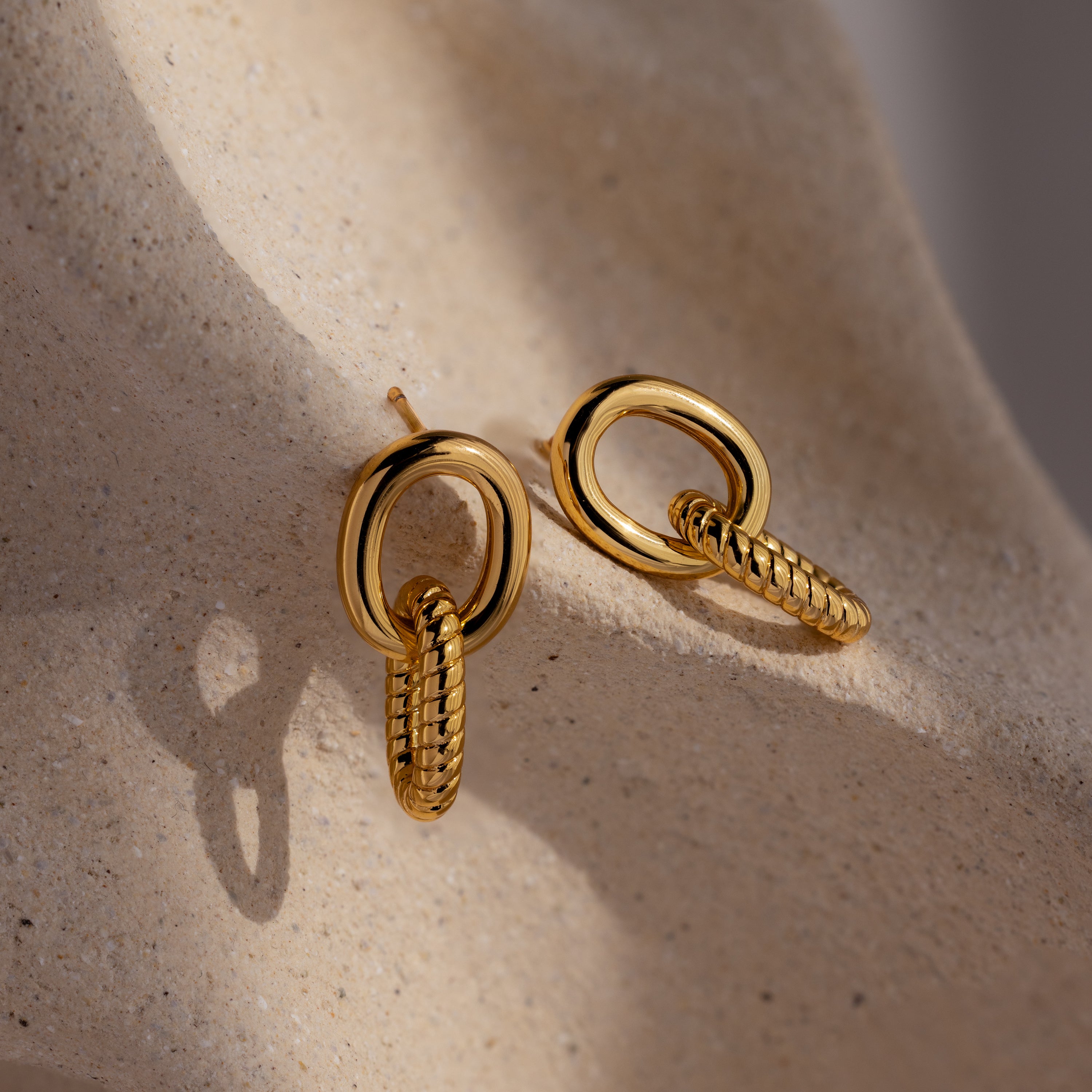 The Twisted Link Earrings in 18K Gold feature interlocking oval and braided loops, elegantly displayed on a textured beige surface.
