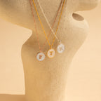 Three Pearl Initial Disc Necklaces engraved with F, T, and R are displayed on a beige sculpture—a timeless personalized set ideal for those who love custom initial necklace styles.