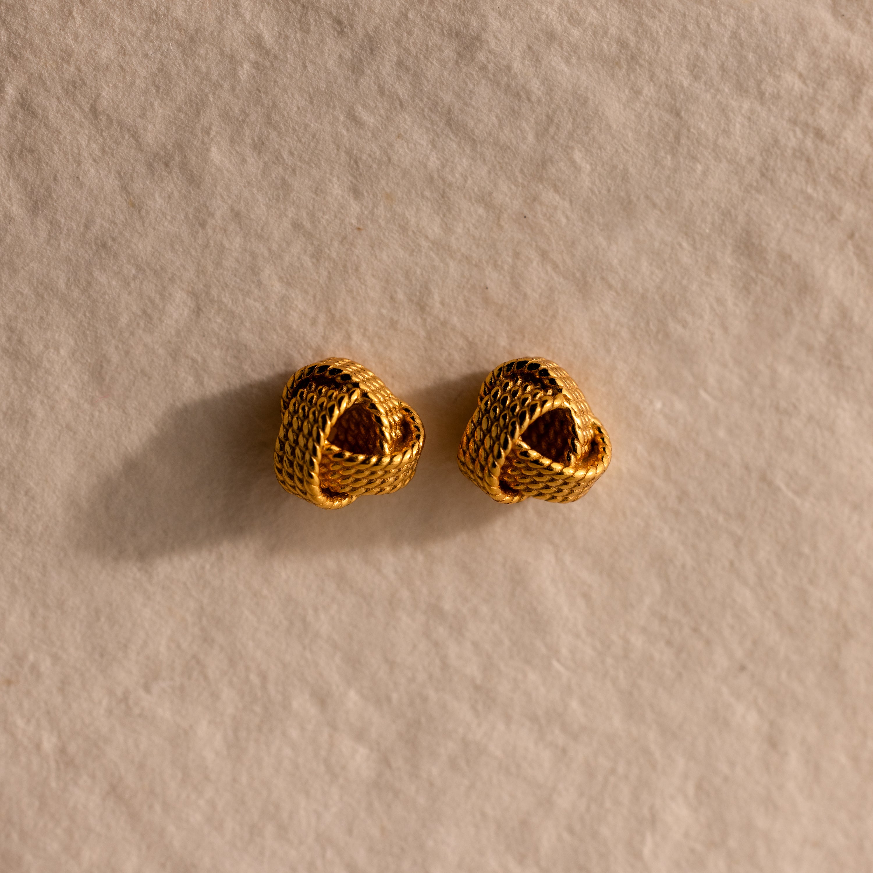 The Rope Knot Studs rest elegantly on a textured beige surface, showcasing timeless style and subtle sophistication.