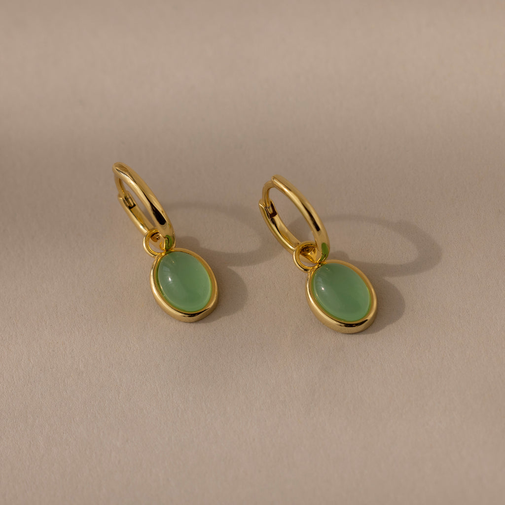 The Jade Drop Hoops are gold huggie earrings featuring oval jade gemstone pendants, shown on a neutral beige background.