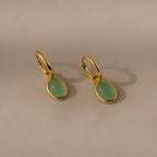 The Jade Drop Hoops are gold huggie earrings featuring oval jade gemstone pendants, shown on a neutral beige background.