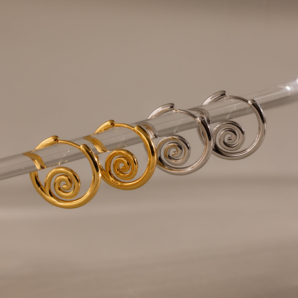 Two pairs of Spiral Swirl Hoops—one gold, one silver—displayed on a clear rod with a neutral backdrop; these earrings add the perfect boho touch to your beach vacation outfits.