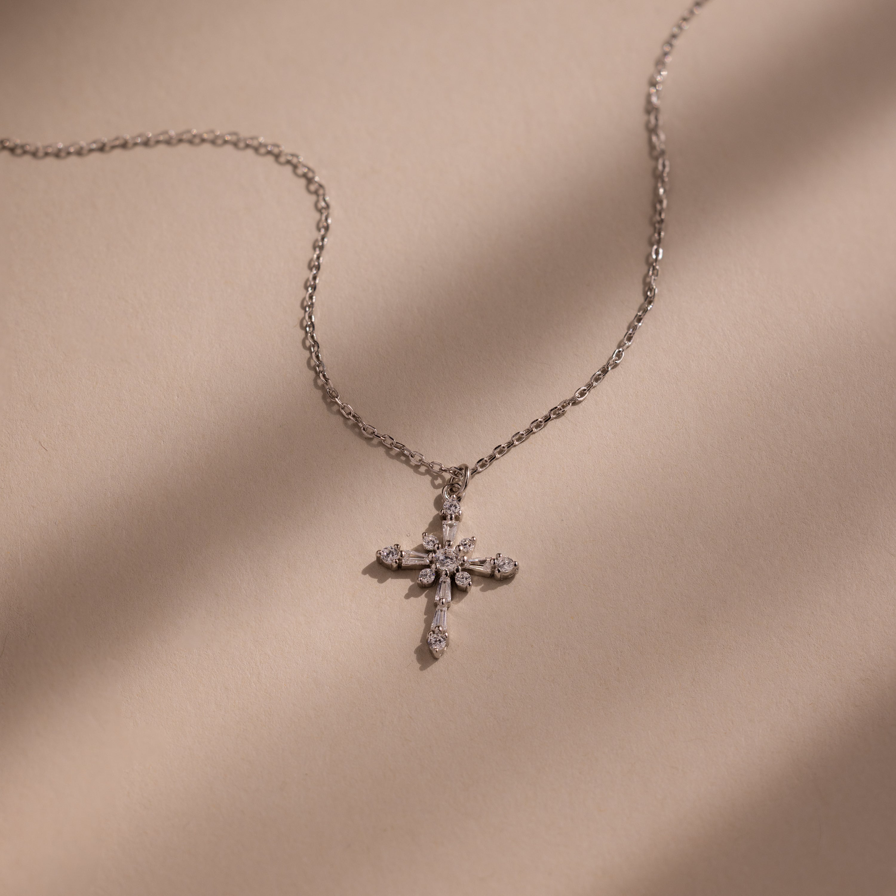 The Vintage Diamond Cross Necklace in Silver is displayed on a beige surface with soft shadow lines.