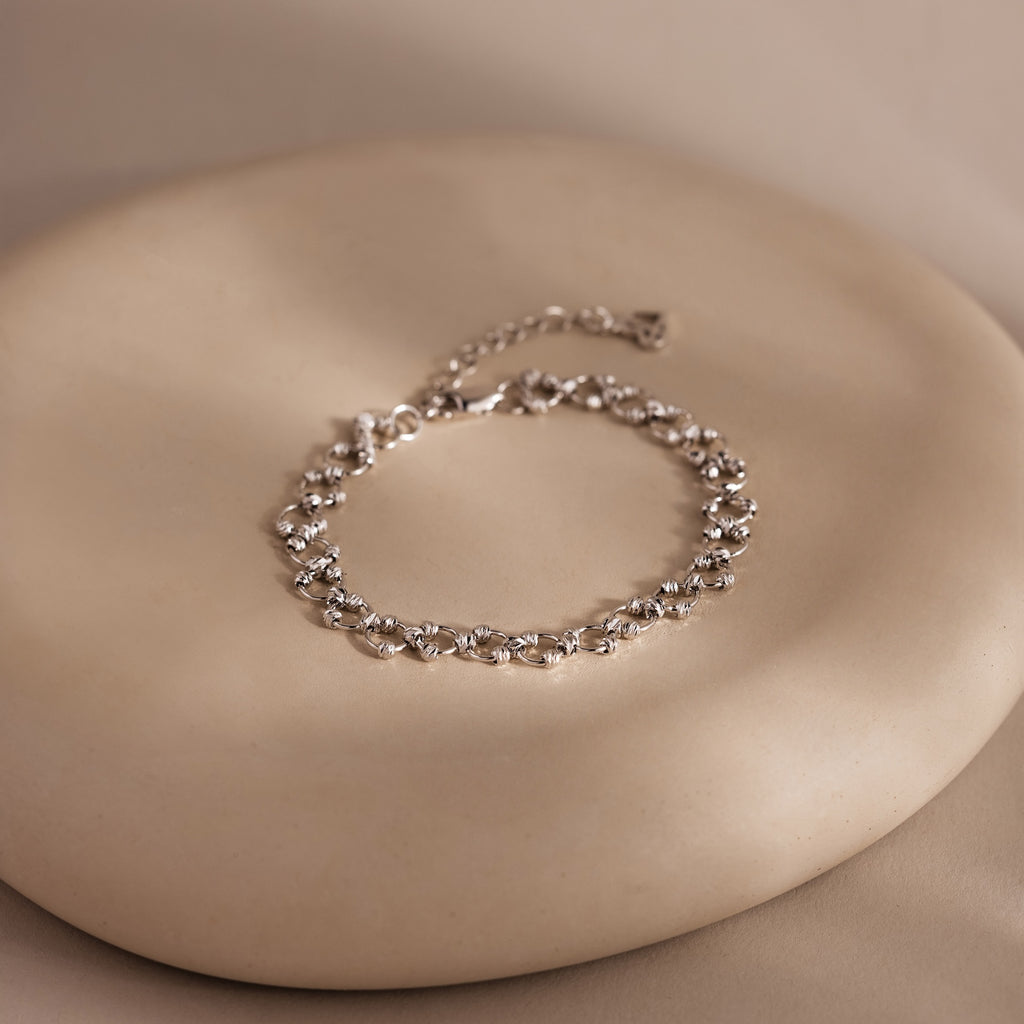 The Orbit Chain Bracelet in Silver, featuring delicate floral links, is showcased on a smooth round beige surface—an exquisite piece of elegant jewelry.