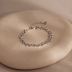The Orbit Chain Bracelet in Silver, featuring delicate floral links, is showcased on a smooth round beige surface—an exquisite piece of elegant jewelry.