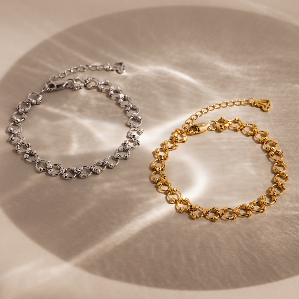 Two elegant Orbit Chain Bracelets—one silver, one gold—are displayed on a beige surface, enhanced by soft light reflections.