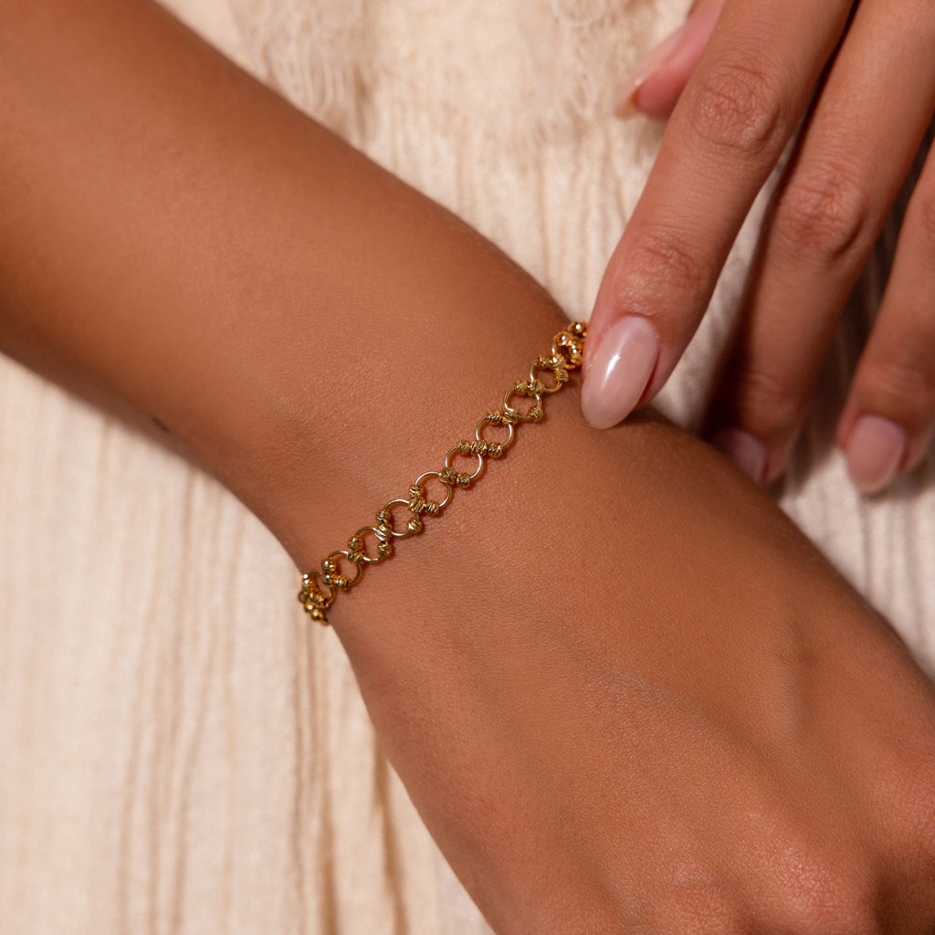 A hand adorned with the Orbit Chain Bracelet, featuring interlocking loops, rests on a cream-colored dress.