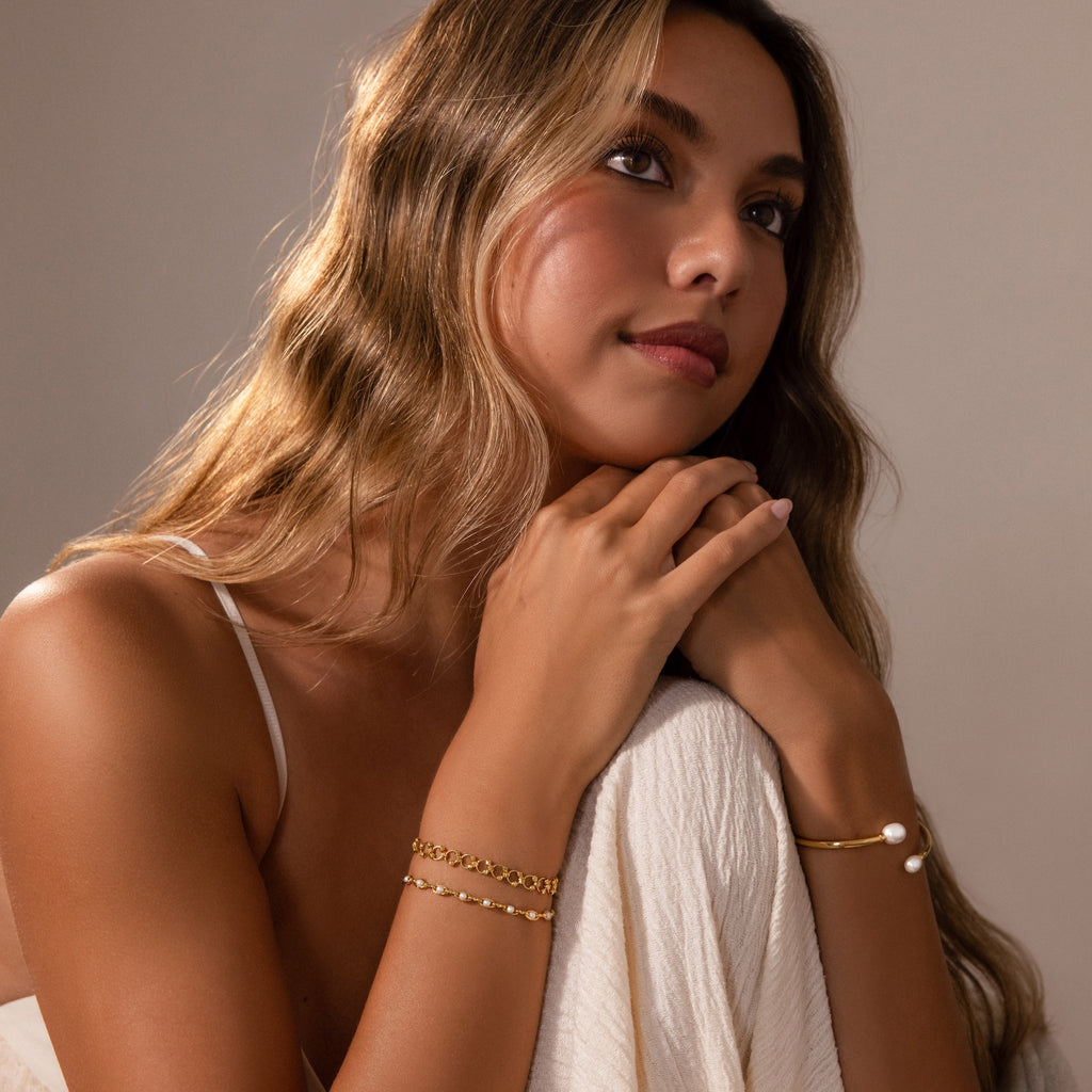 Woman with wavy hair wears the Orbit Chain Bracelet and gold bangles, resting her chin on her hands while gazing thoughtfully upward.