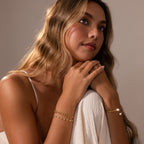 Woman with wavy hair wears the Orbit Chain Bracelet and gold bangles, resting her chin on her hands while gazing thoughtfully upward.
