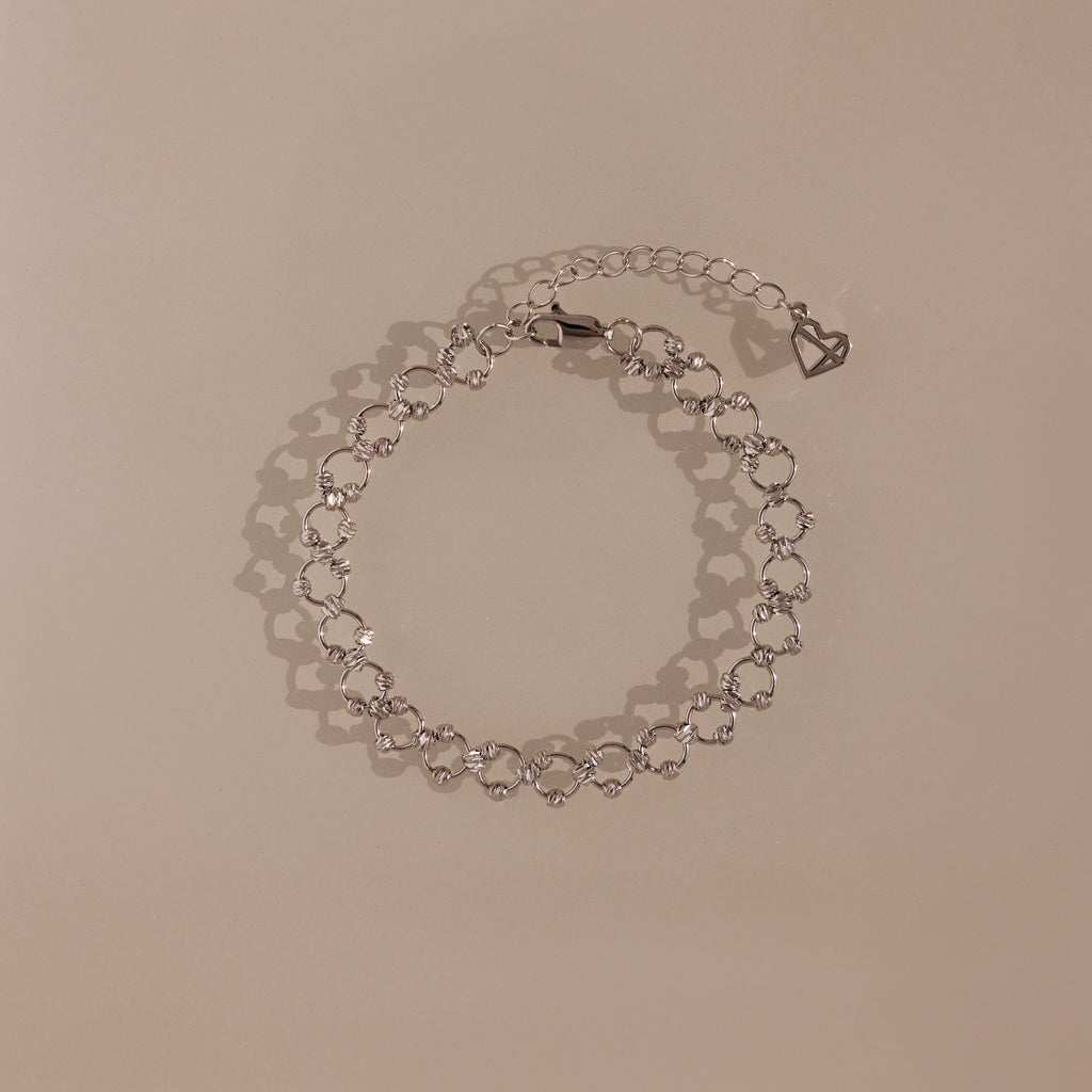 The Orbit Chain Bracelet in Silver features delicate heart-shaped links and an adjustable chain, showcased on a beige background—ideal for fans of elegant jewelry.