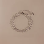 The Orbit Chain Bracelet in Silver features delicate heart-shaped links and an adjustable chain, showcased on a beige background—ideal for fans of elegant jewelry.