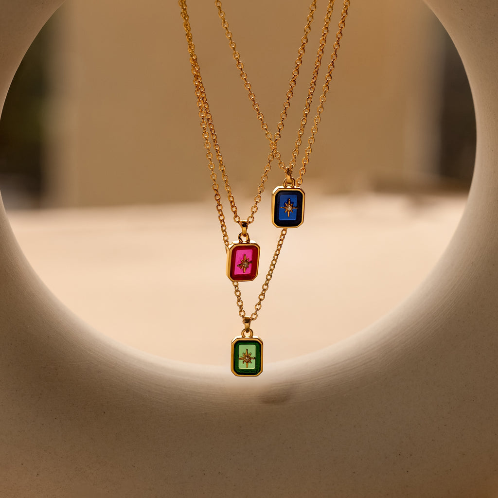 Three North Star Birthstone Necklaces with gold chains and rectangular crystal pendants in pink, blue, and green, each adorned with a diamond-set North Star design.