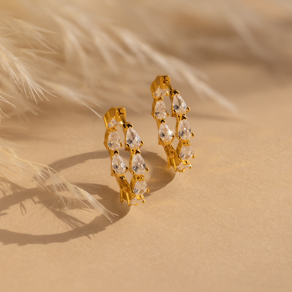 The Inside Out Pear Diamond Hoops feature gold hoops embellished with multiple teardrop-shaped CZ diamonds, showcased on a beige surface alongside soft dried pampas grass.