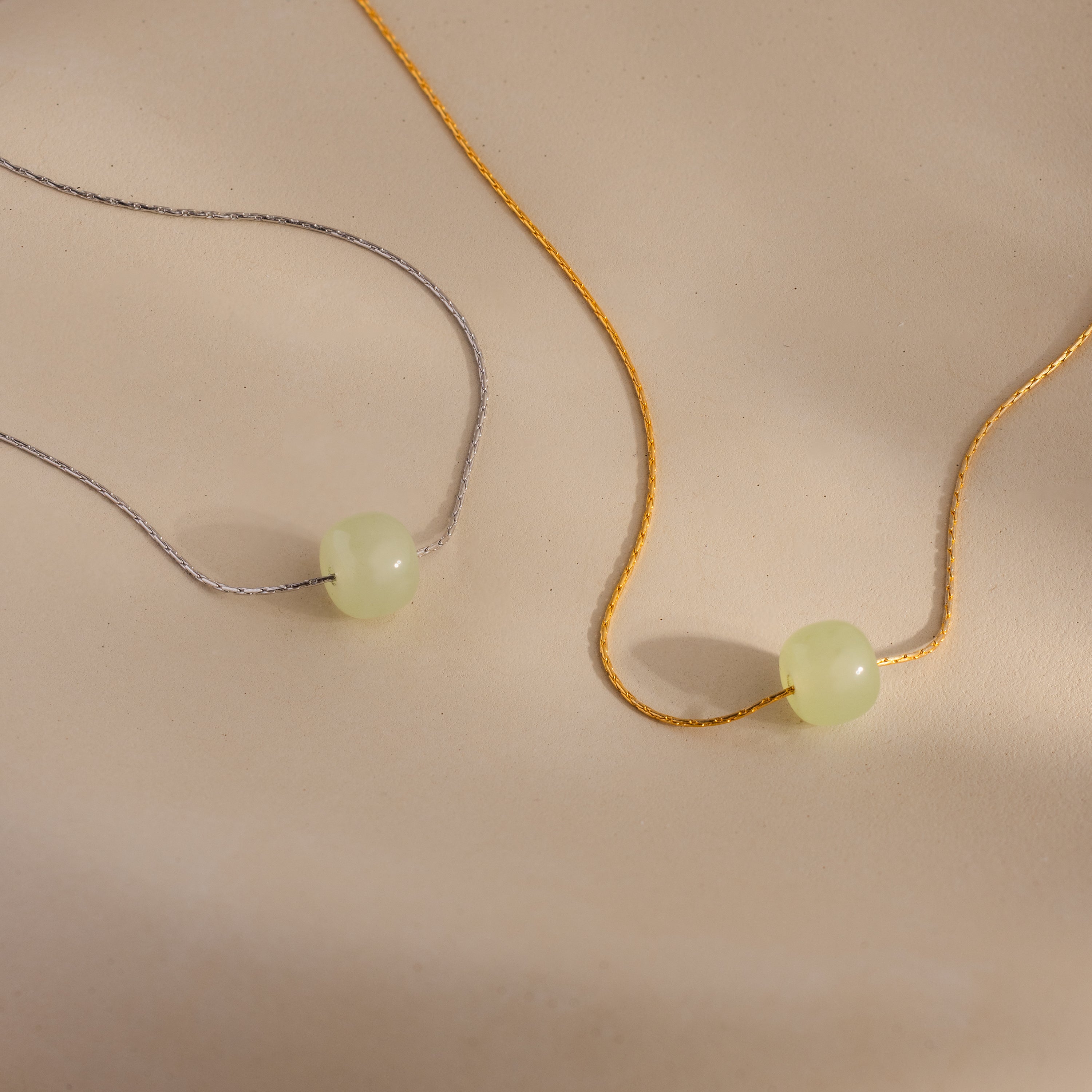 Close-up of a two dainty chain necklaces in gold and silver, each featuring a single round green jade bead, displayed against a neutral surface.