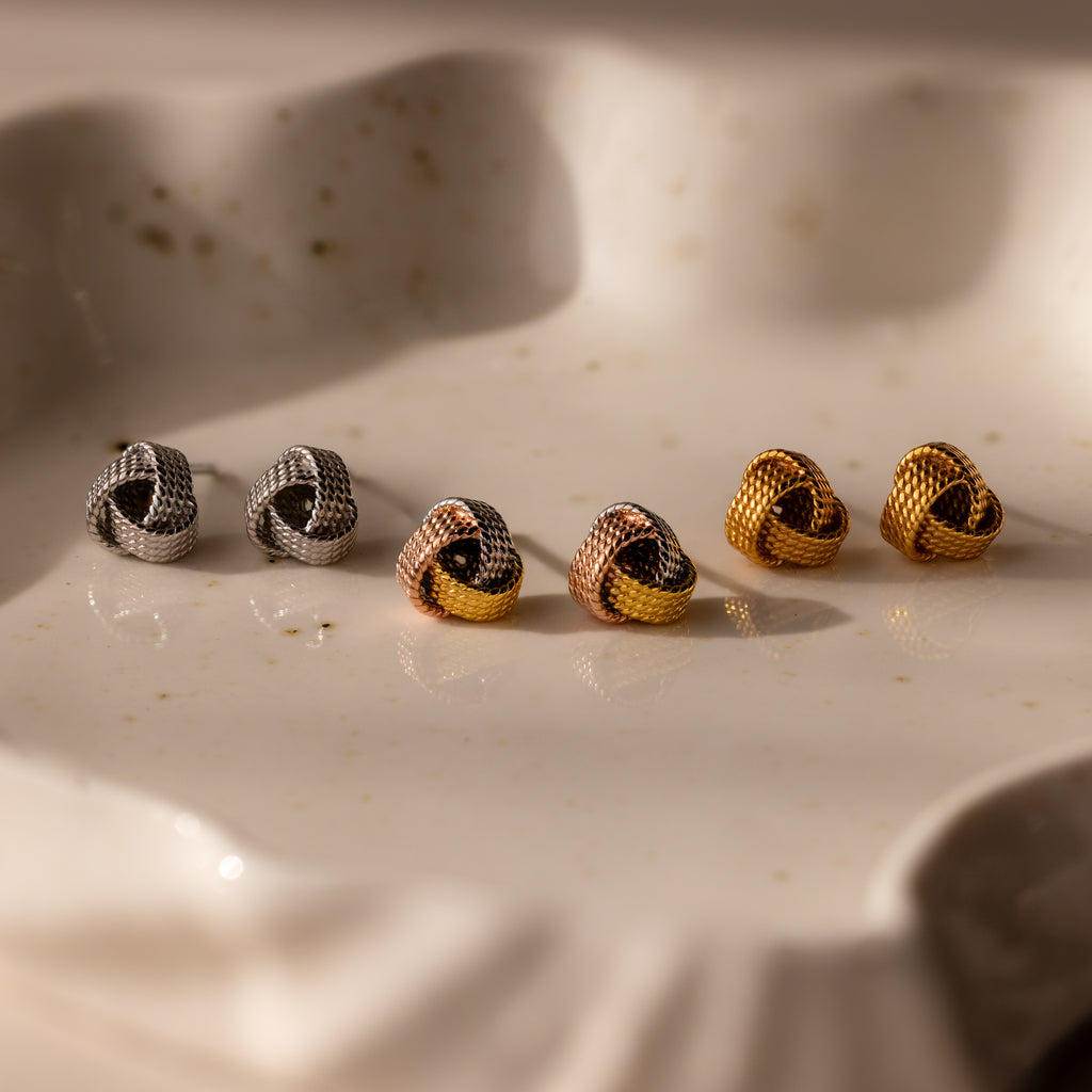 The Rope Knot Studs set includes three pairs of knot-shaped earrings in silver, rose gold, and gold, elegantly displayed on a white ceramic dish—an ideal choice for adding timeless style to any look.