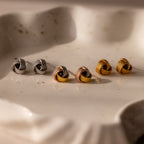 The Rope Knot Studs set includes three pairs of knot-shaped earrings in silver, rose gold, and gold, elegantly displayed on a white ceramic dish—an ideal choice for adding timeless style to any look.