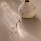 The Turquoise Hand Chain Bracelet in Silver is elegantly displayed draped over a white ceramic vase set on a beige surface.