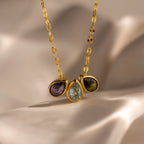 The Multiple Teardrop Birthstone Necklace features three gemstones—purple, green, and blue—on a gold chain against a soft beige background. A personalized jewelry piece and thoughtful gift.