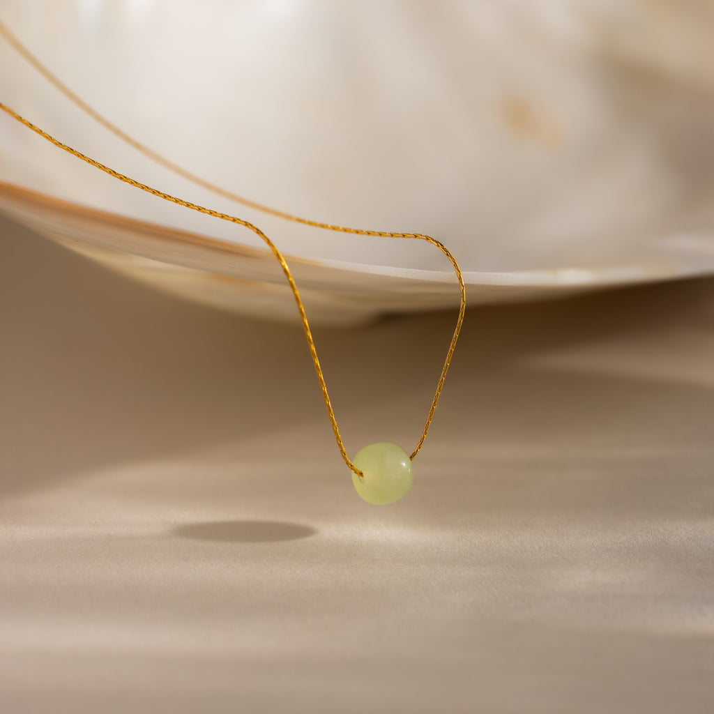 Close-up of a delicate gold chain necklace featuring a single round green jade bead, draped from a white shell dish.